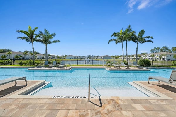 Resort swimming pool with shallow and deep sections, palm trees, lounge chairs, and waterfront homes beyond the railing under clear blue sky
